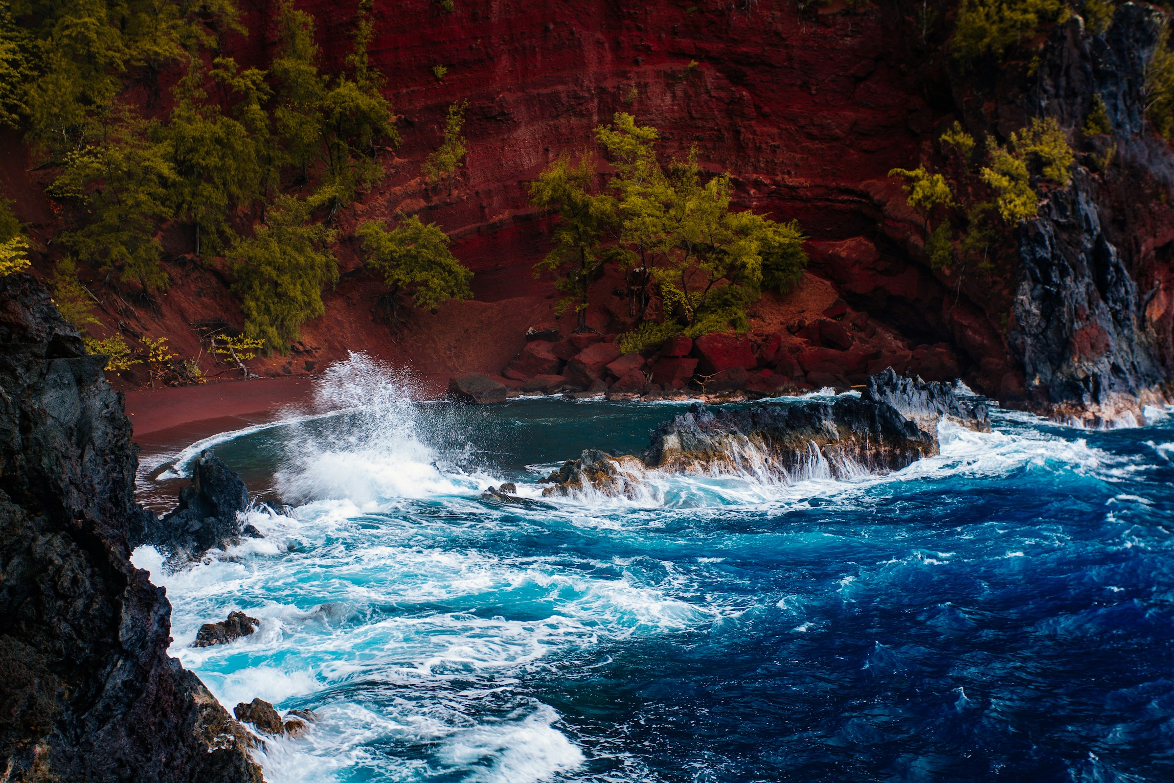 Waves crashing against a red sand beach with lush trees on a rugged coastline.